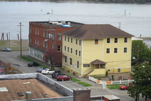 A view of two buildings by a river, one yellow and one red, with parked cars and a cloudy sky.