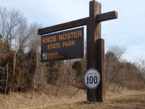 Sign for Knob Noster State Park in Missouri, featuring park name and state logo, surrounded by bare trees.