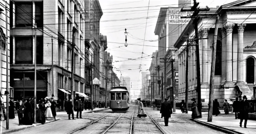 Black and white photo of a busy city street with a tram, pedestrians, and buildings lining the road.