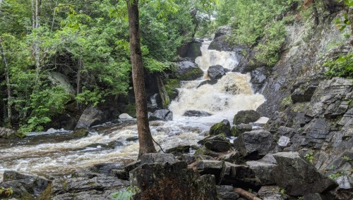 A cascading waterfall flows over rocky terrain, surrounded by lush green trees and foliage.