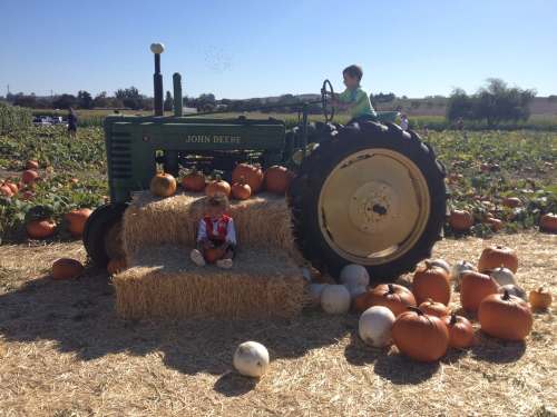 Two children play on a John Deere tractor surrounded by pumpkins and hay bales in a sunny pumpkin patch.
