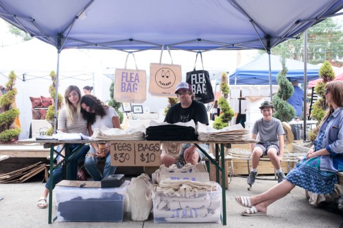 A market stall with people selling bags and tees, surrounded by greenery and other vendors under a tent.