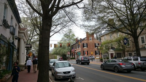 A street scene with shops, cars parked along the curb, and trees lining the road under a cloudy sky.