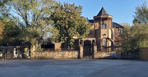 A large, unique brown house with a turret, surrounded by trees and a stone wall, set against a clear blue sky.