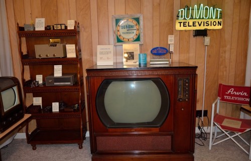 Vintage wooden television set displayed in a retro room with shelves of old electronics and a neon DuMont sign.