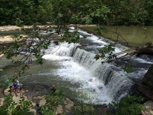 A serene waterfall cascades over rocks, surrounded by lush greenery and people enjoying the natural scenery.