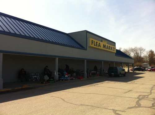 Exterior of a flea market building with a large sign, featuring people and items for sale outside.