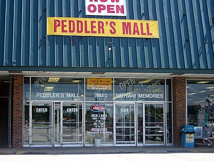 Exterior of Peddler's Mall with large "Now Open" sign and glass doors, showcasing a welcoming storefront.
