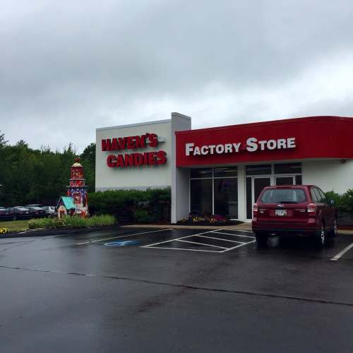 Exterior of Maven's Candies Factory Store, featuring a sign and a parked car, surrounded by greenery and cloudy skies.