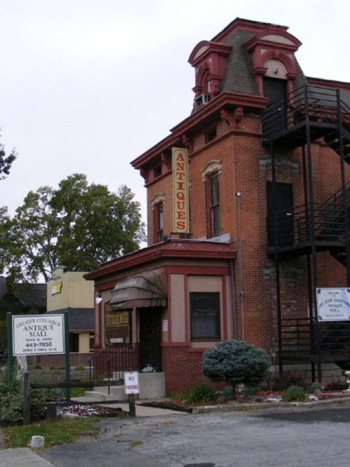 A vintage brick building with a sign reading "Antiques," featuring a staircase and surrounding greenery.