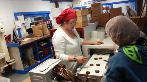 A chocolatier in a red headscarf prepares chocolates while a child watches in a colorful kitchen.