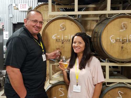 Two people stand in front of wooden barrels, smiling and holding glasses of beer in a brewery setting.