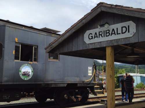 A vintage train at Garibaldi station, with a sign overhead and a scenic backdrop of trees and hills.