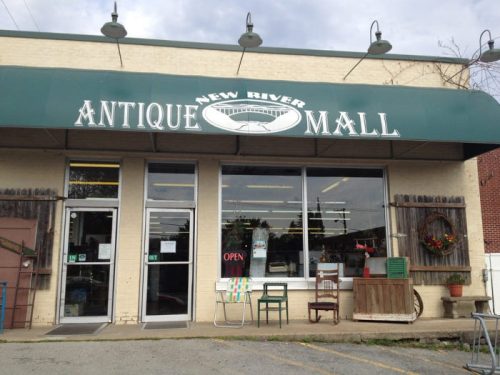 Exterior of the New River Antique Mall, featuring a green awning and an "Open" sign, with various items displayed outside.