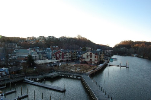A serene waterfront view featuring buildings along a river, with docks and hills in the background under a clear sky.