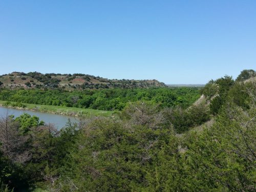 A scenic view of a river surrounded by lush greenery and rolling hills under a clear blue sky.