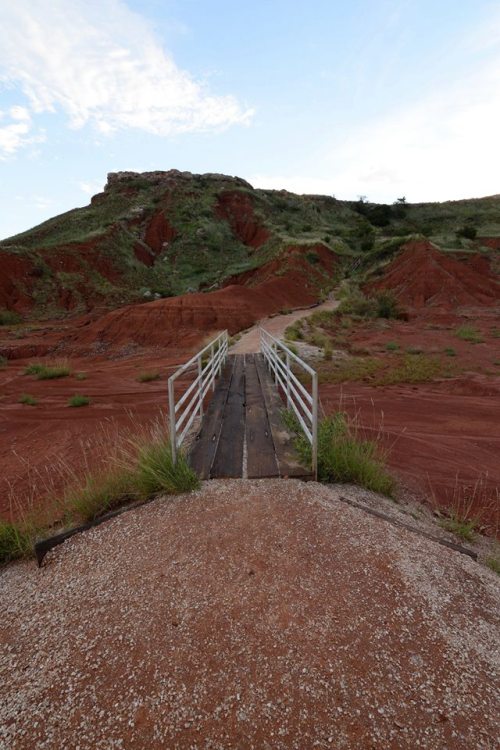 A wooden bridge leads over a red, rocky landscape with grassy patches and a hill in the background under a blue sky.