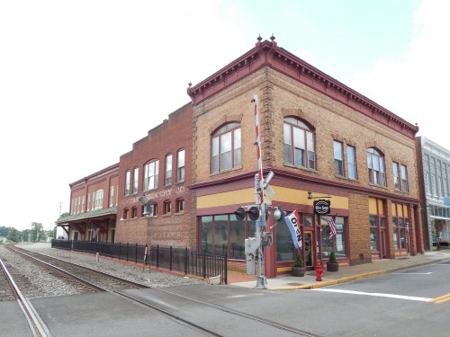 Historic brick building with large windows, featuring a "Open" sign, next to railroad tracks.