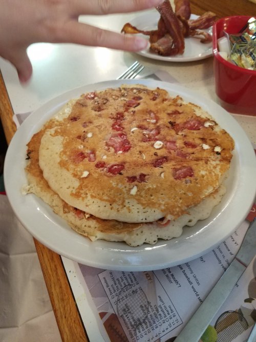 A stack of pancakes with strawberries, served on a white plate, with a hand reaching into the frame.