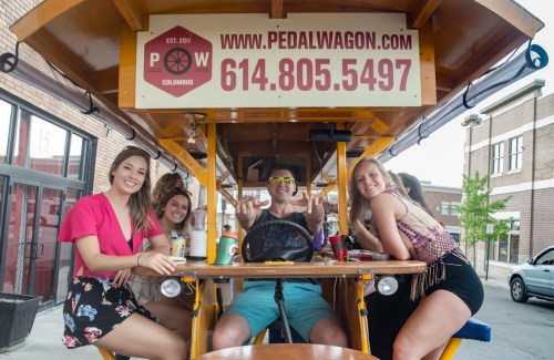 A group of four friends enjoying drinks on a pedal wagon, smiling and posing for the camera.
