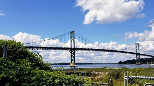 A suspension bridge spans a river under a blue sky with fluffy clouds, surrounded by greenery and water.