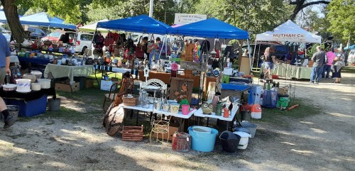 A bustling outdoor market with various stalls, tables filled with items, and blue tents providing shade.