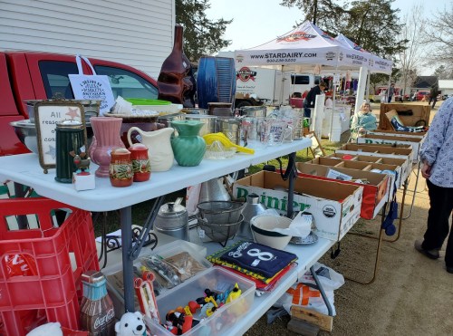 A table filled with various vintage items and collectibles at an outdoor market, with tents in the background.