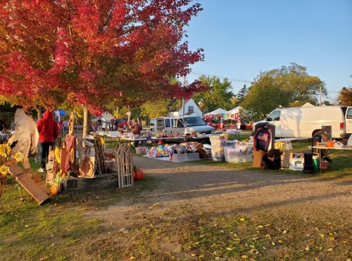 A vibrant outdoor market scene with colorful fall foliage, vendors, and various items for sale under a clear blue sky.