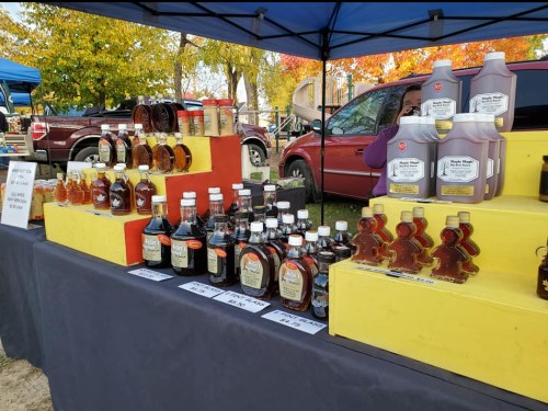 A market stall displays various bottles of maple syrup in different sizes, with colorful signage and autumn foliage in the background.