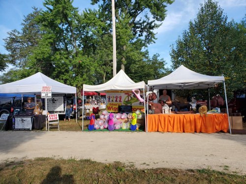 Two vendor tents at an outdoor market, one selling wigs and the other featuring various items, with trees in the background.