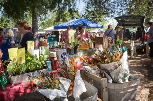 A bustling outdoor market with tables filled with flowers, plants, and various goods, surrounded by shoppers and tents.