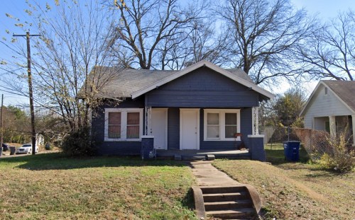 A small blue house with a front porch, surrounded by trees and grass, on a clear day.