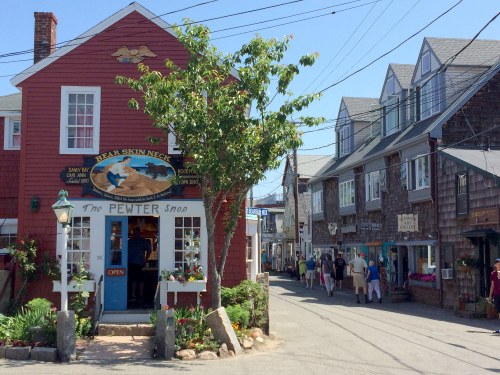 A quaint street scene featuring a red shop, "The Pewter Shop," and charming buildings with shoppers strolling by.