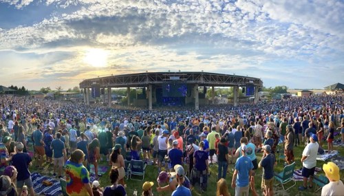 A large crowd gathers at an outdoor concert venue under a colorful sky at sunset.