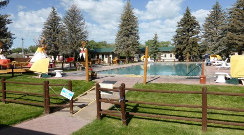 A sunny pool area surrounded by trees, with tipis, lounge chairs, and a grassy lawn.