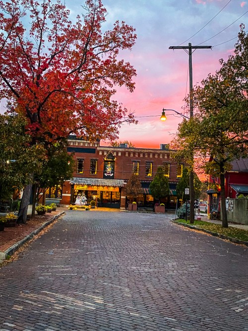 A cobblestone street lined with trees, vibrant autumn leaves, and a colorful sunset behind a brick building.
