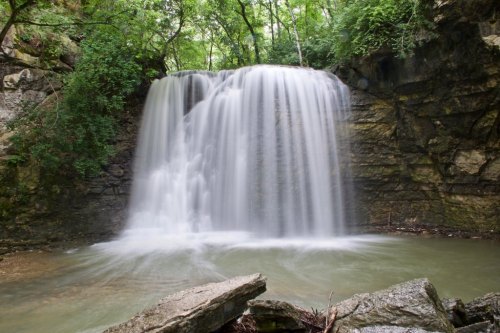 A serene waterfall cascades over rocky cliffs into a calm pool, surrounded by lush greenery.
