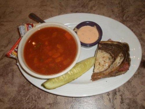 A plate with soup, a sandwich, a pickle, and a small dish of sauce.