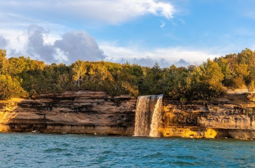 A waterfall cascades over rocky cliffs into a serene blue lake, surrounded by lush green trees under a partly cloudy sky.