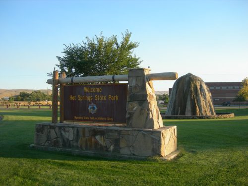 Welcome sign for Hot Springs State Park, featuring a stone structure and a large rock, set in a grassy area.