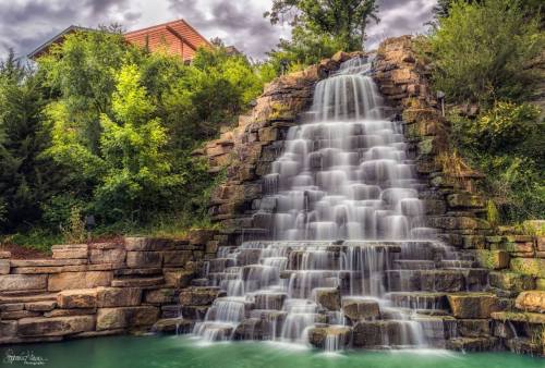 A serene waterfall cascades over stone steps into a tranquil pool, surrounded by lush greenery and a cloudy sky.