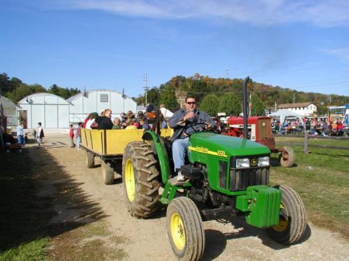 A man drives a green tractor pulling a yellow wagon filled with people on a sunny day at a farm event.