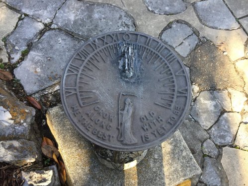 A weathered sundial on a stone path, with inscriptions and a small figure at the base.