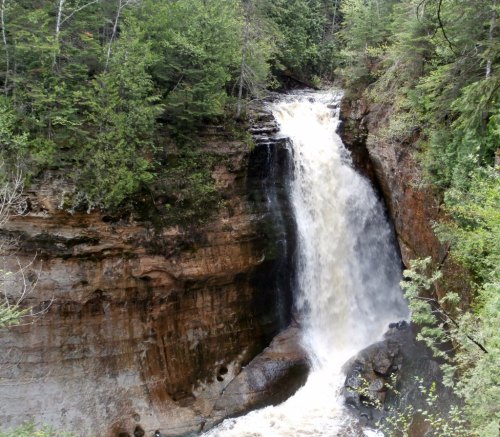 A cascading waterfall flows over rocky cliffs, surrounded by lush green trees and foliage.