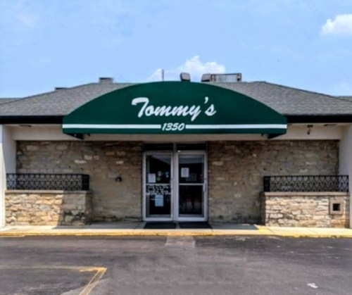 A storefront with a green awning reading "Tommy's" and a stone facade, set against a clear blue sky.