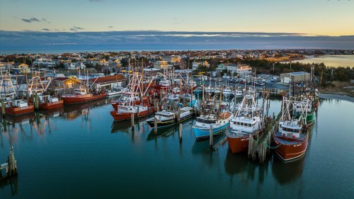 Aerial view of a fishing harbor at sunset, featuring several boats docked in calm waters with a town in the background.