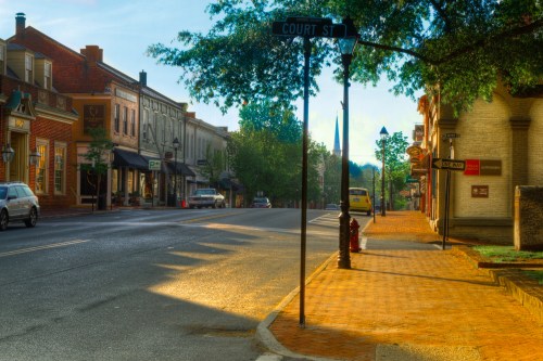 A quiet street scene with shops lining the road, a street sign for Court St, and trees casting shadows in the sunlight.