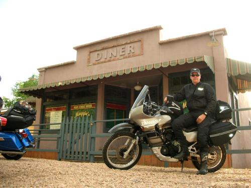 A motorcyclist in black gear sits on a bike in front of a vintage diner with a sign reading "Diner."