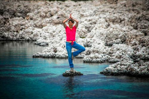 A person in a pink shirt and jeans stands on one leg on a rock in calm water, surrounded by rocky terrain.