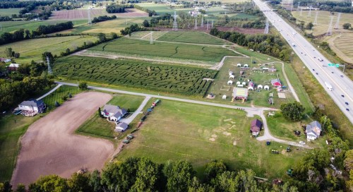 Aerial view of a rural area with fields, a maze, and tents near a highway, surrounded by trees and farmland.